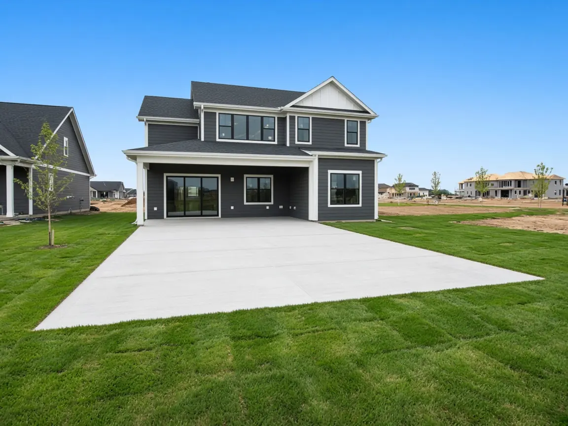 Large new concrete patio behind a newly built charcoal two-story home with fresh sod and homes under construction in the distance
